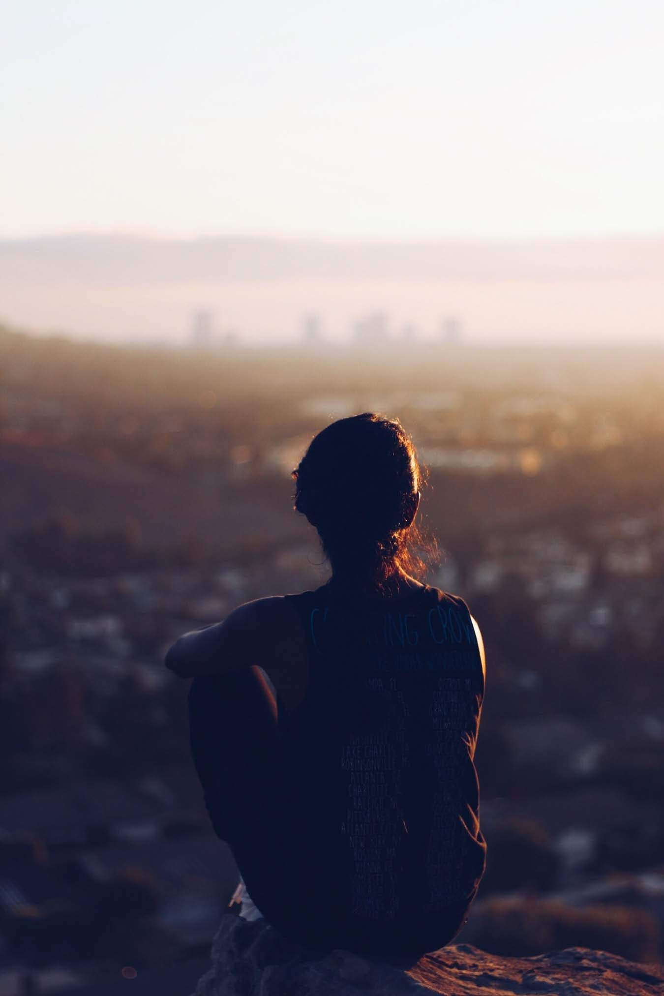 Eine junge Frau von hinten zu sehen, bei Sonnenuntergang, auf einem Stein sitzend während sie auf eine unscharfe Stadt am Horizont blickt