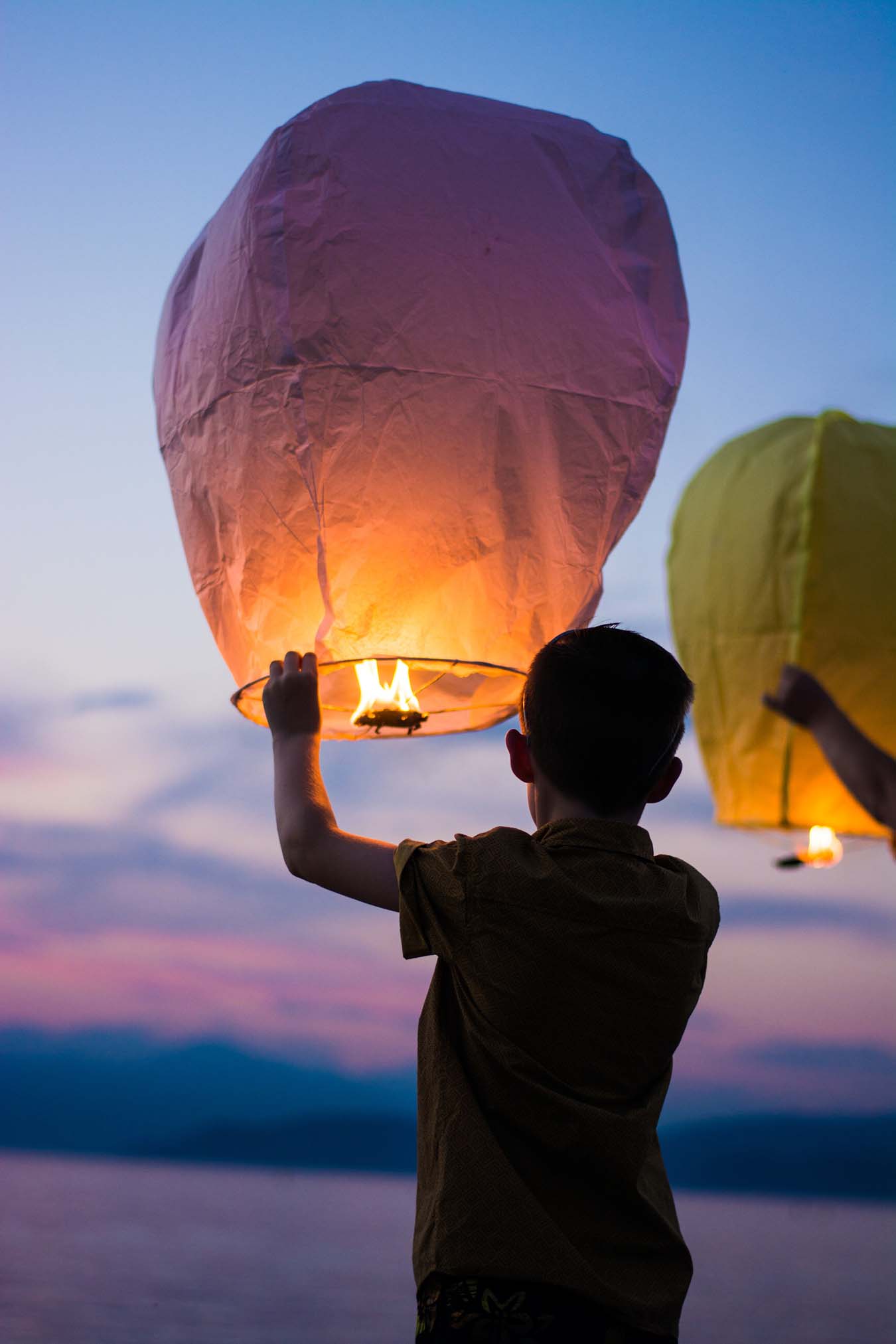 a boy seen from behind at dusk starting to let fly a yellow flame driven paper balloon