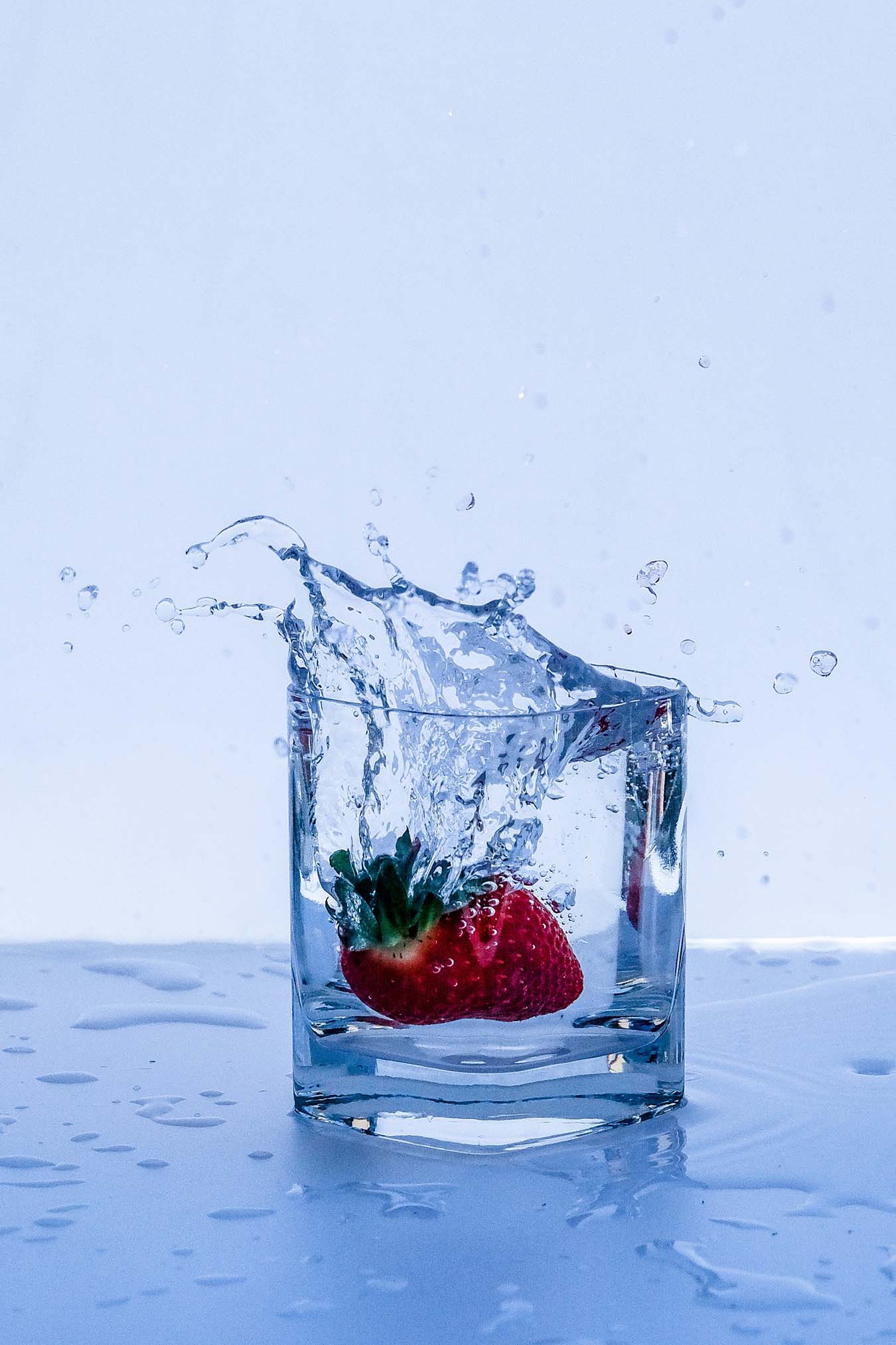 In ein volles Wasserglas fällt eine rote Erdbeere. Es spritzt Wasser heraus.