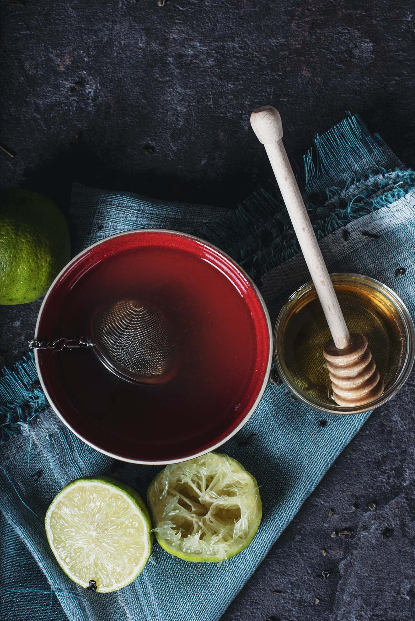 On a blue cloth stands a red cup with a tea infuser, a jar with honey and two squeezed limes.