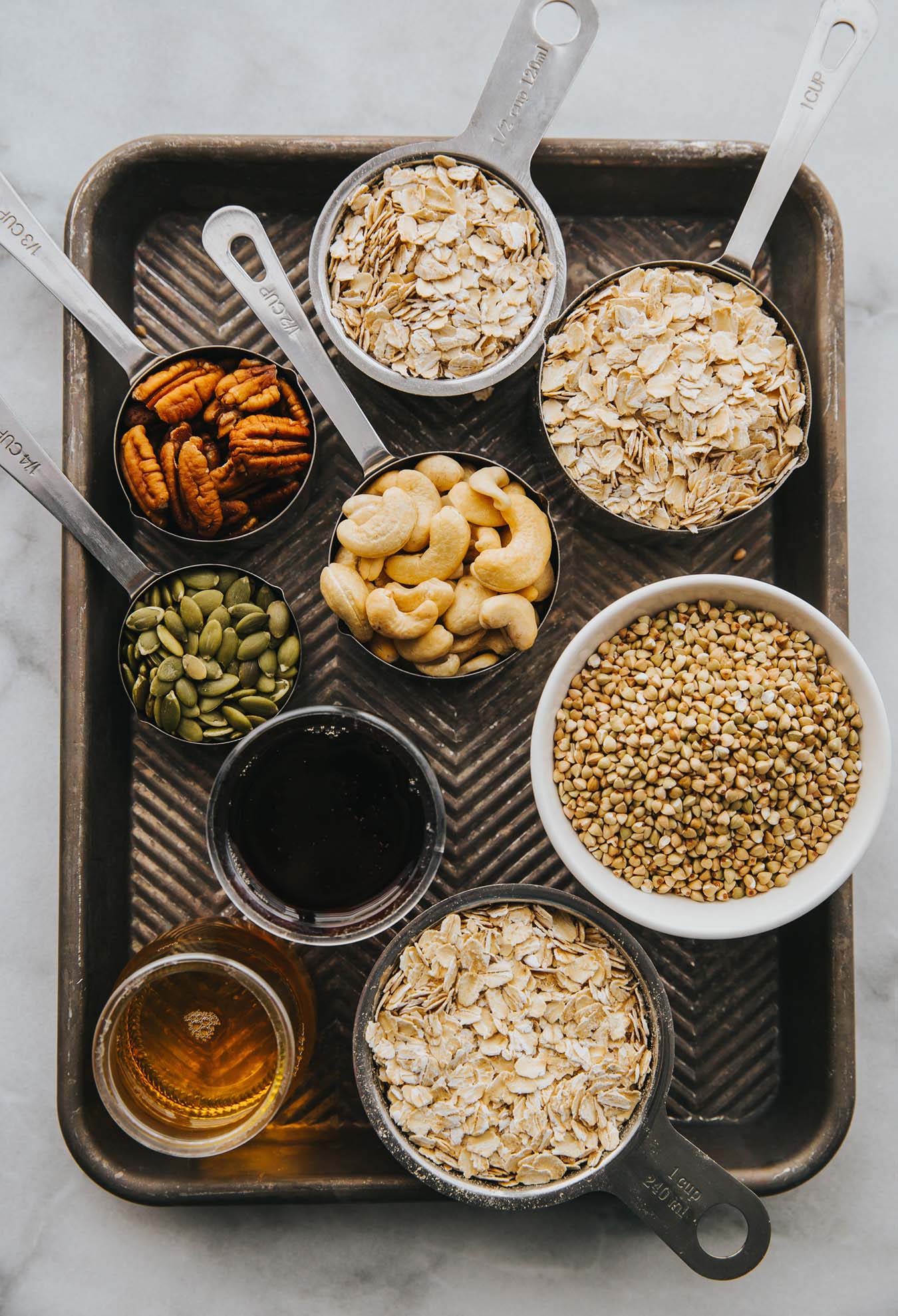 Auf einem alten Backblech stehen kleine Schüsselchen mit verschiedenen Zutaten wie Cashews, Buchweizen, Haferflocken, Pekannüsse und Kürbiskerne.