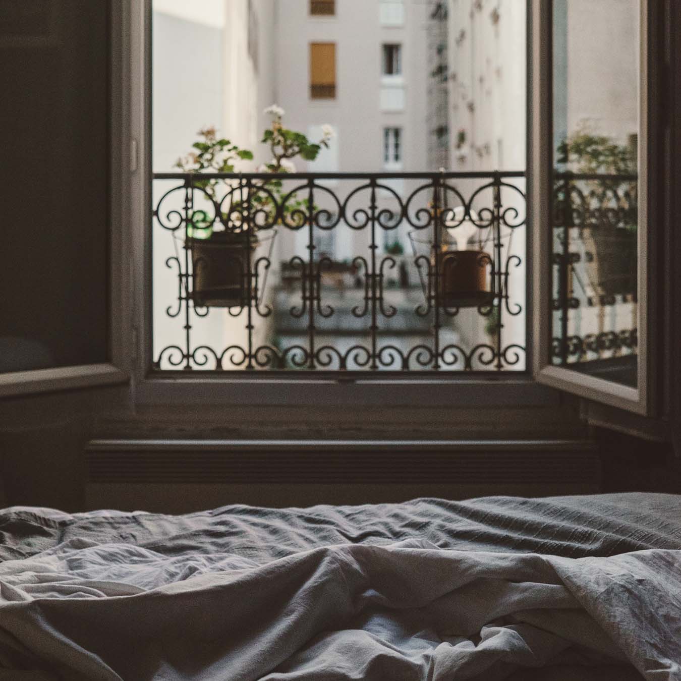 A made bed stands in front of an open window with flowers.