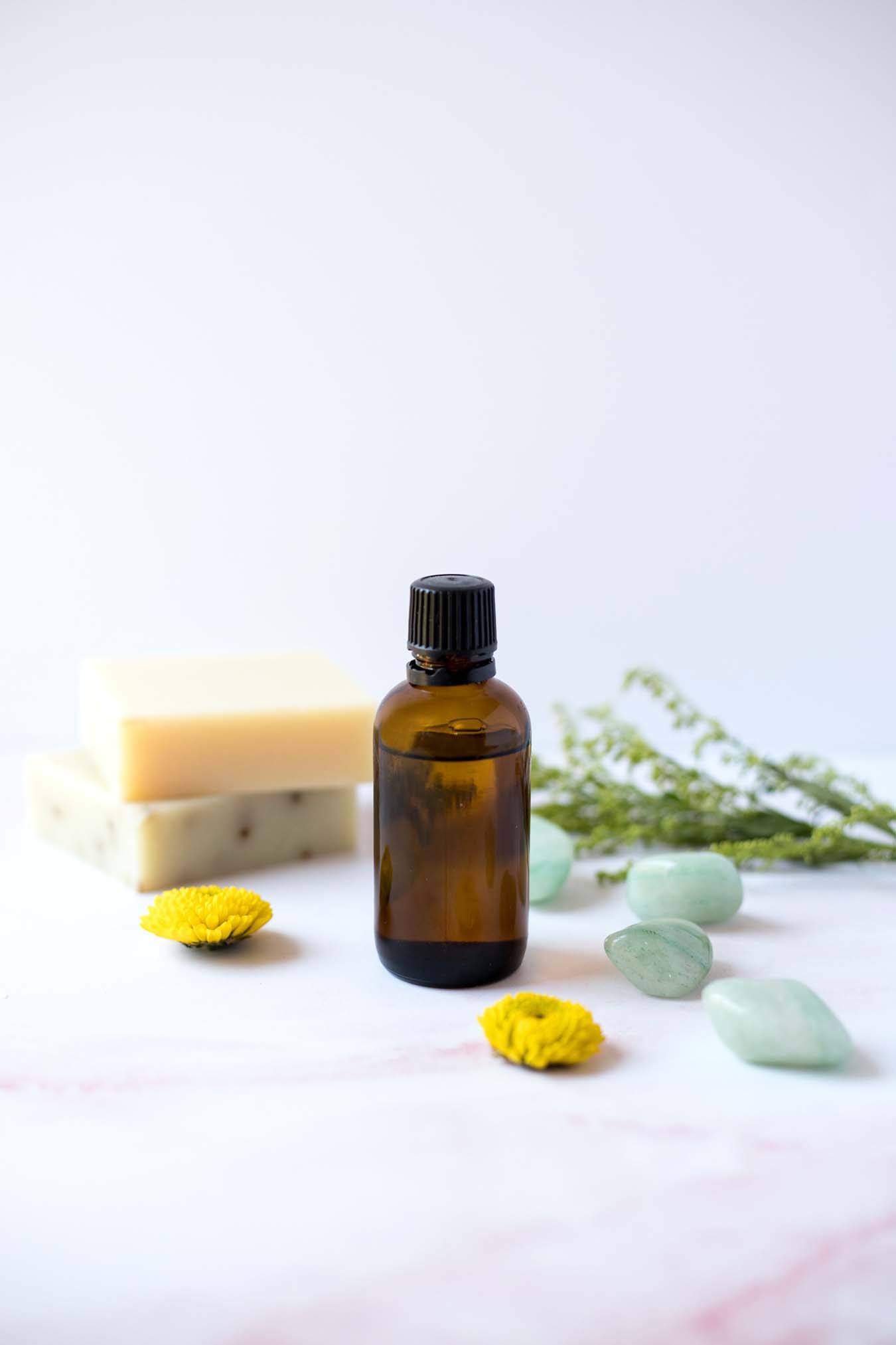 A brown glas bottle filled with essential oils stands on a table that is decorated with blue stones and yellow flowers.