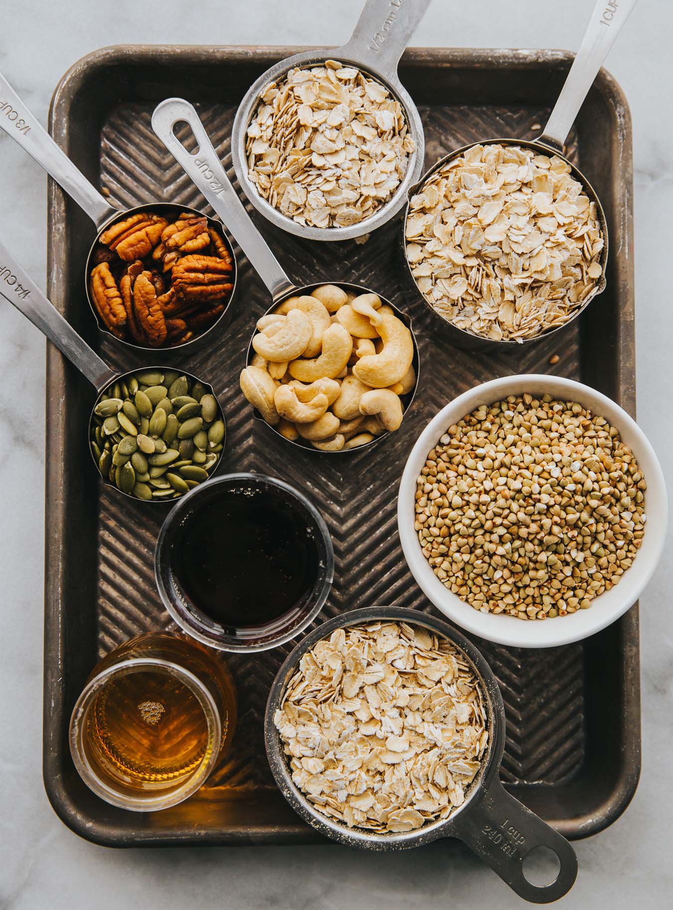 Auf einem alten Backblech stehen kleine Schüsselchen mit verschiedenen Zutaten wie Cashews, Buchweizen, Haferflocken, Pekannüsse und Kürbiskerne.